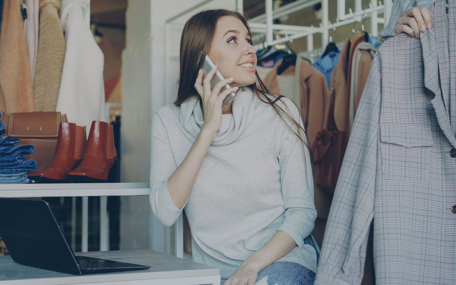 A woman on the phone in a clothing shop, with a laptop open on the counter beside her