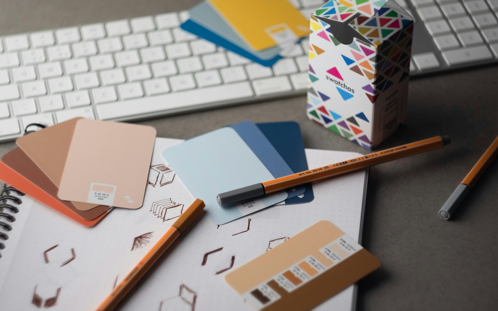 Paint color swatch cards, pencils, and hand-drawn sketches spread across a desk beside a keyboard