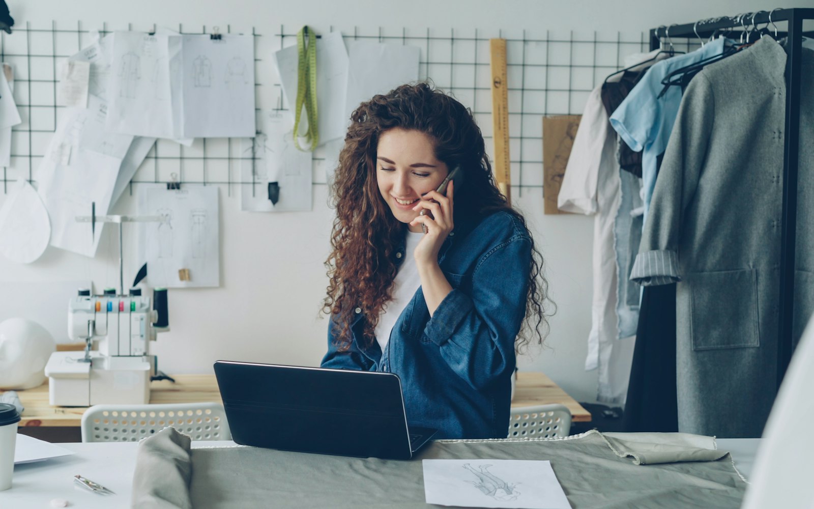 A woman on the phone at a laptop in a sewing studio, with clothing racks and pattern sketches behind her