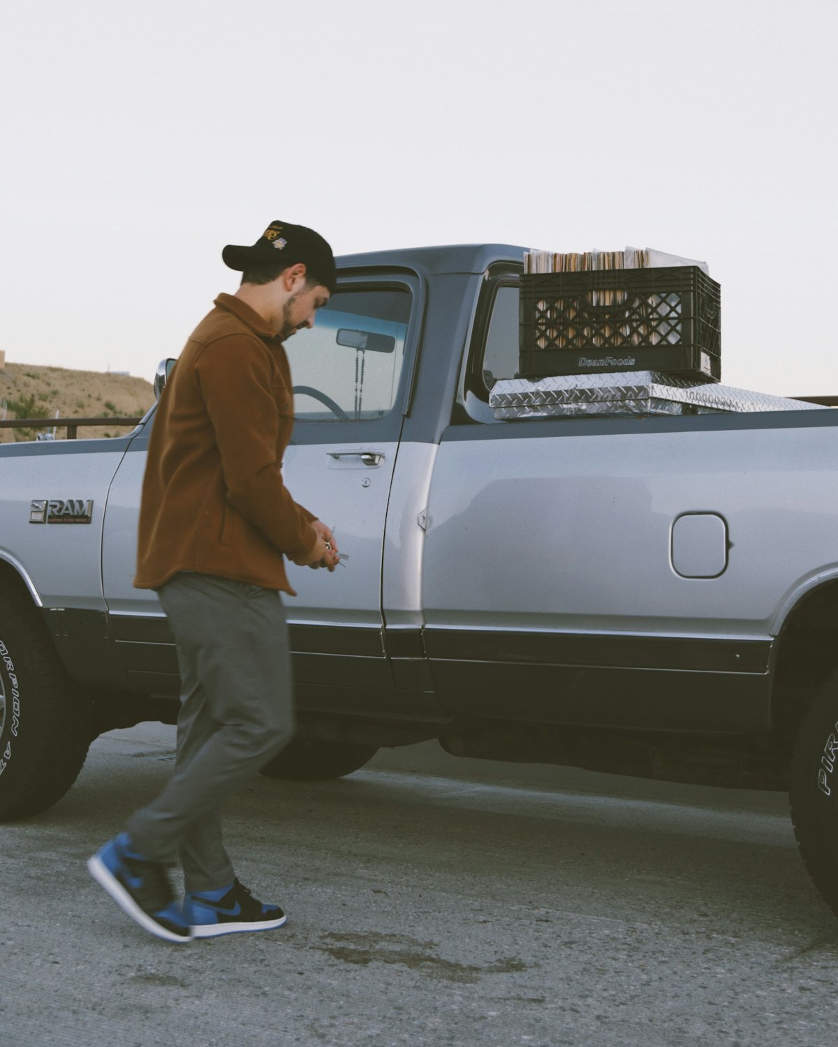 A man in a brown jacket and cap walking beside a silver pickup truck, looking down at his hands