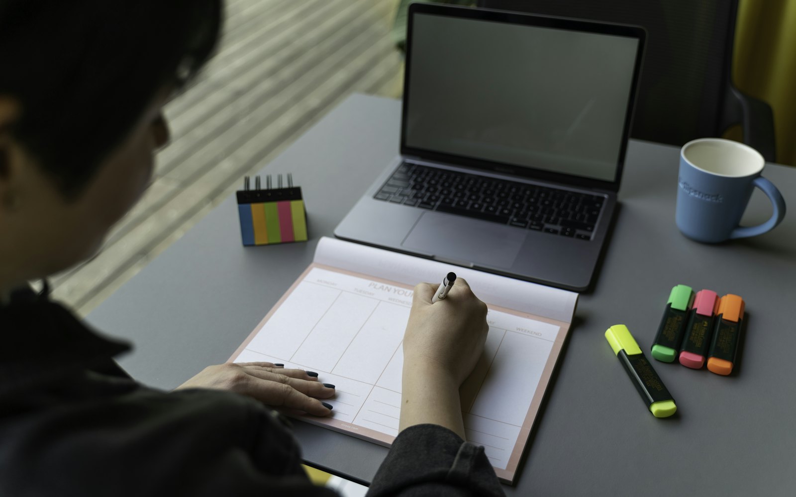 A person writing in a weekly planner at a desk, with a laptop, sticky notes and a mug beside them
