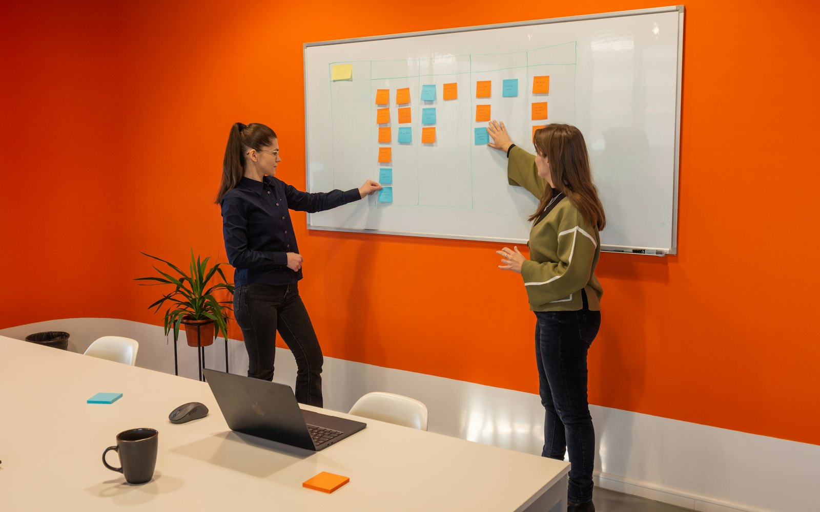 Two people placing sticky notes on a whiteboard in a meeting room with an orange wall