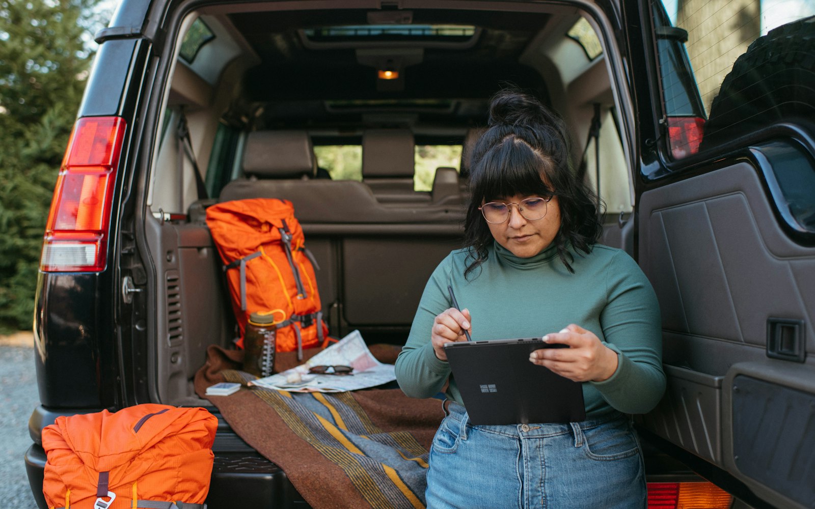 A woman sitting at the open back of an SUV, working on a tablet with a pen