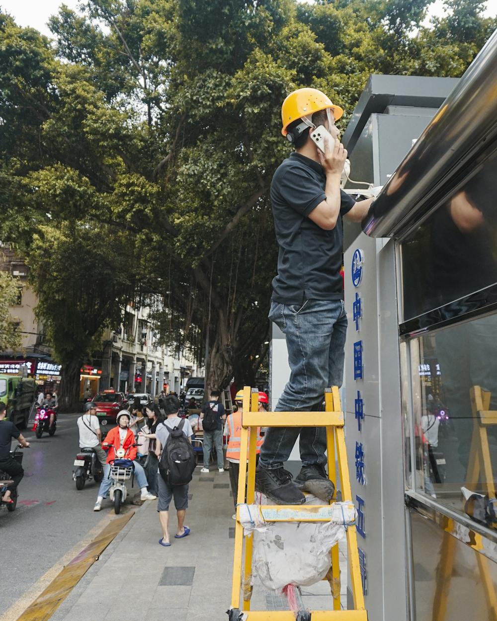 A tradesperson in a yellow hard hat on a ladder, taking a call