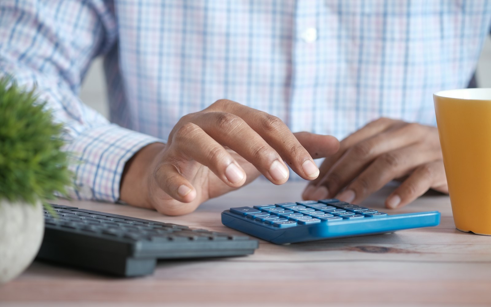 A person's hands using a blue calculator at a wooden desk, with a keyboard, a plant and a mug nearby