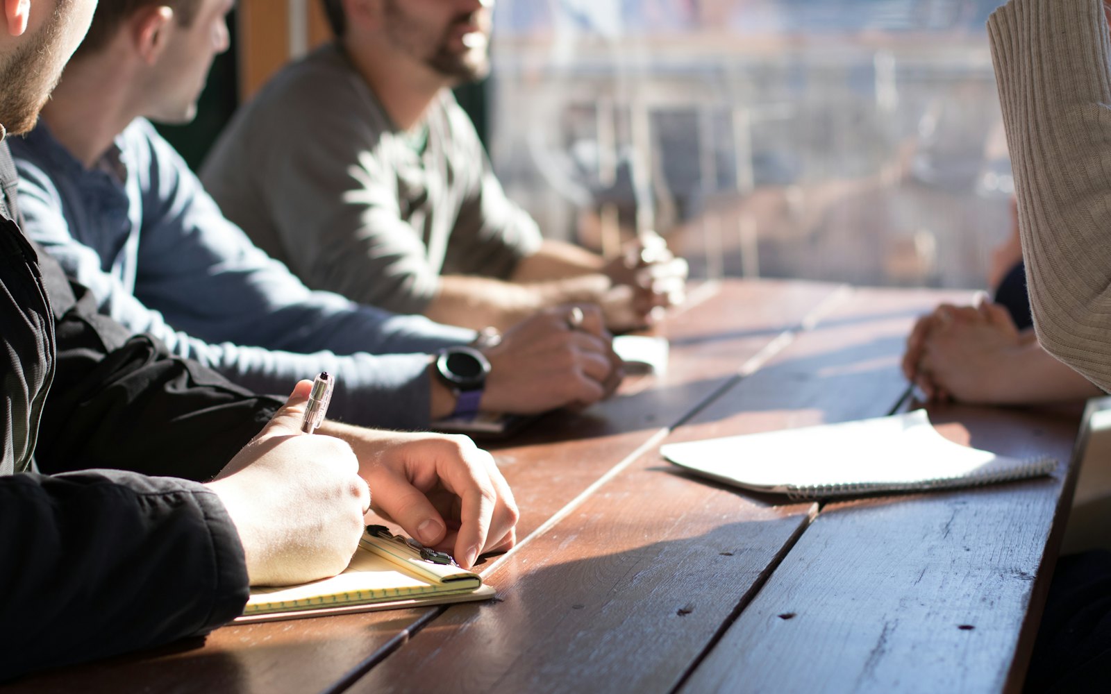 People sitting at a wooden table holding pens and notebooks during a meeting