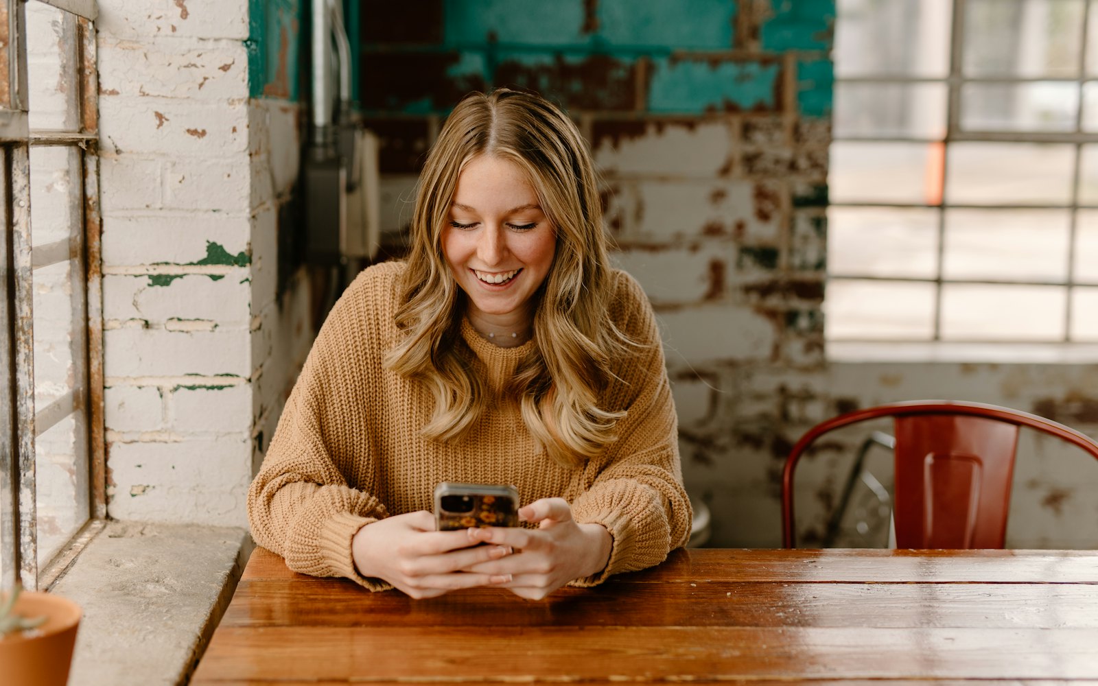 A smiling woman in a tan sweater looks at her phone at a wooden table in a room with painted brick walls