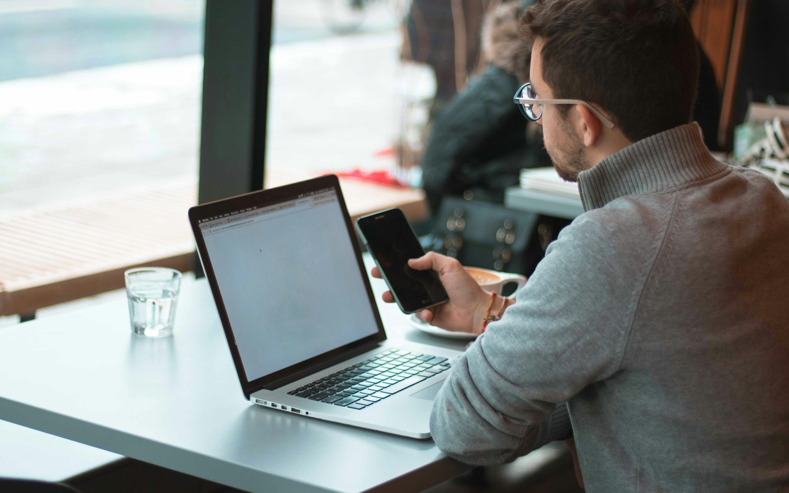A man at a table by a window with a laptop and smartphone