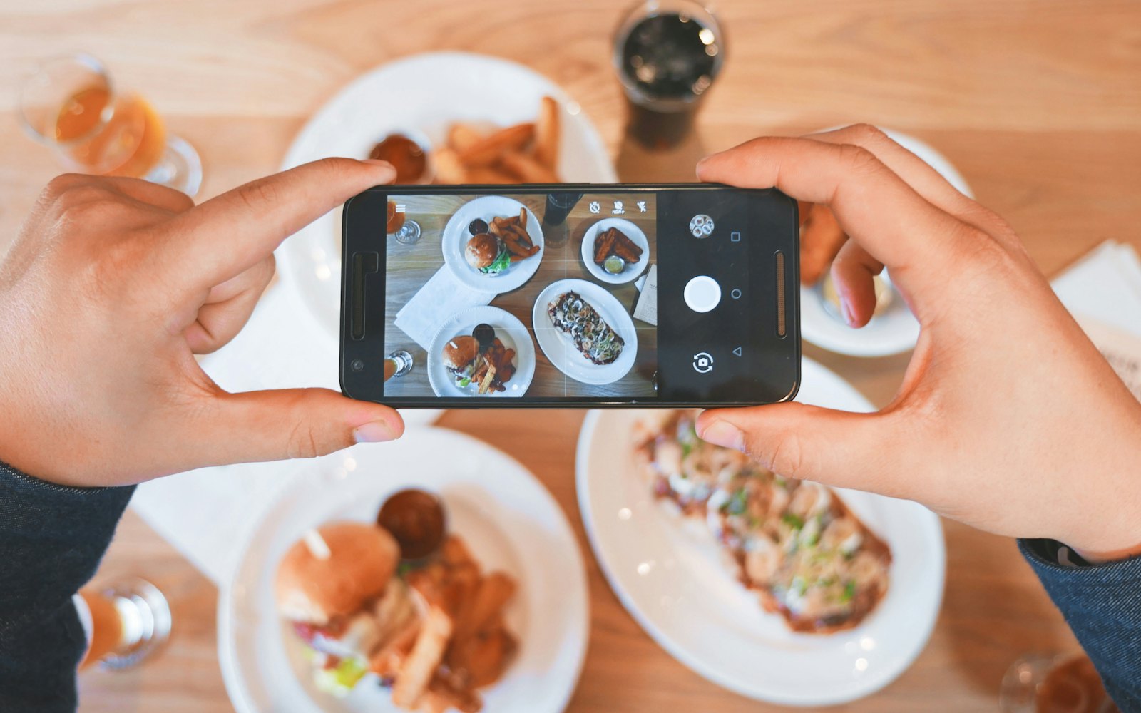 Two hands holding a phone above a restaurant table, photographing burgers, fries, and drinks