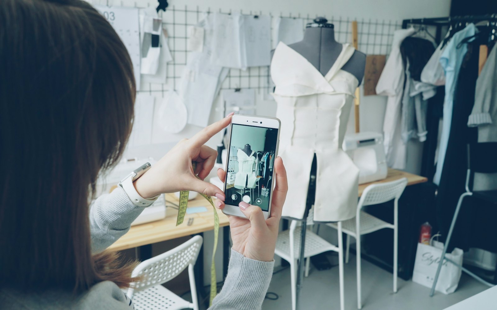 A woman in a sewing studio photographing a dress form with her phone