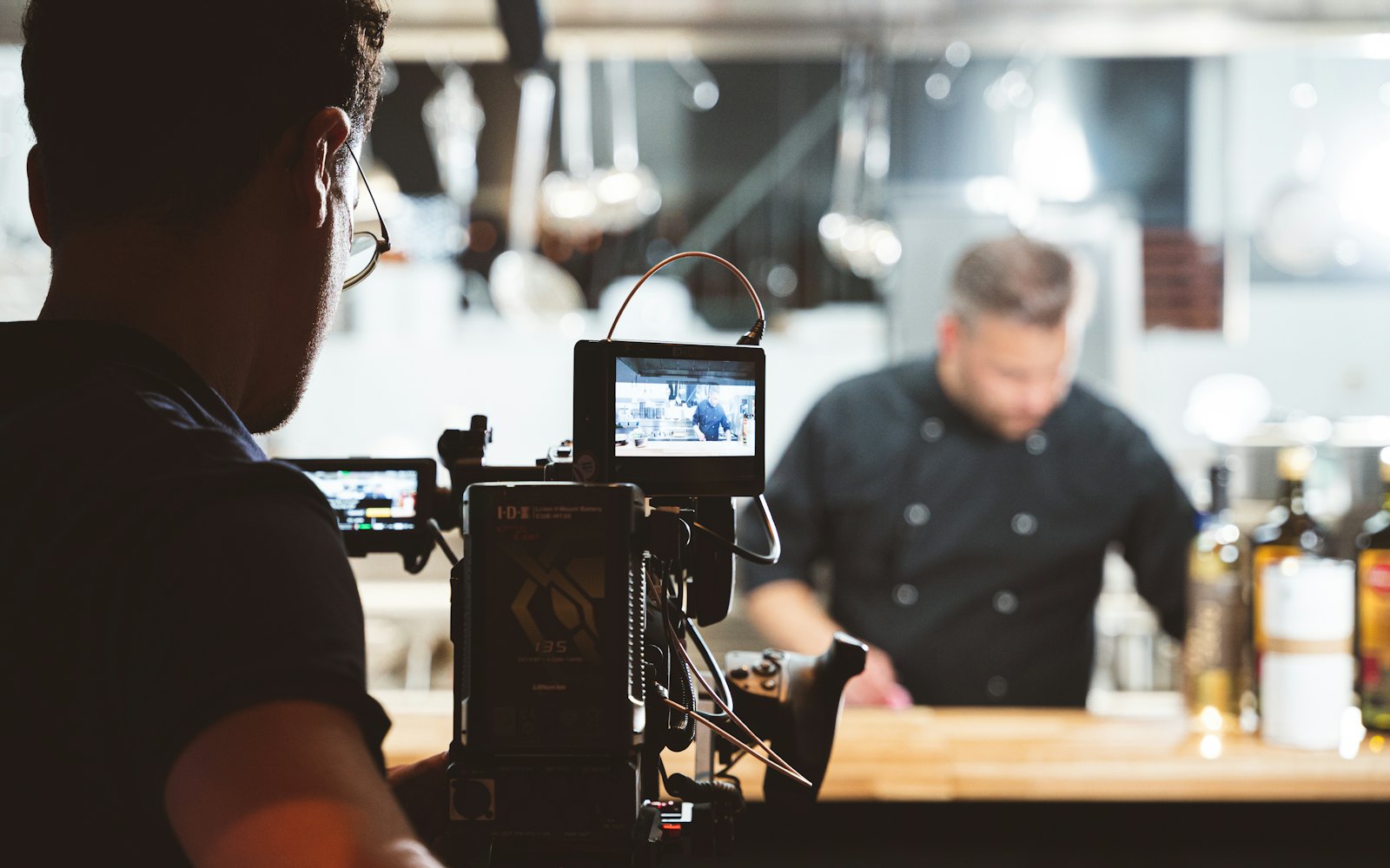 A camera operator filming a chef at work behind a restaurant counter, with the shot showing on the camera monitor