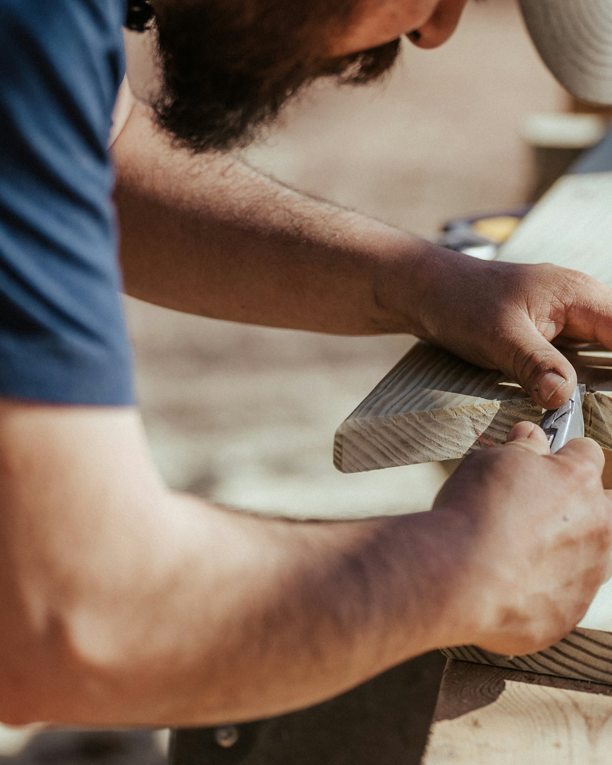 A man in a blue T-shirt working the edge of a wooden board by hand at an outdoor workbench