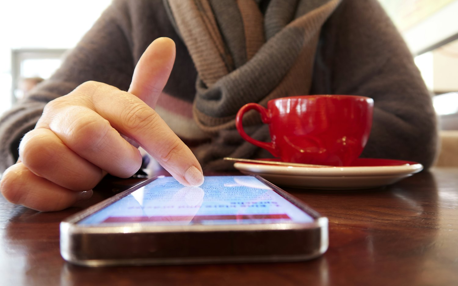 A hand tapping a smartphone screen on a cafe table next to a red coffee cup